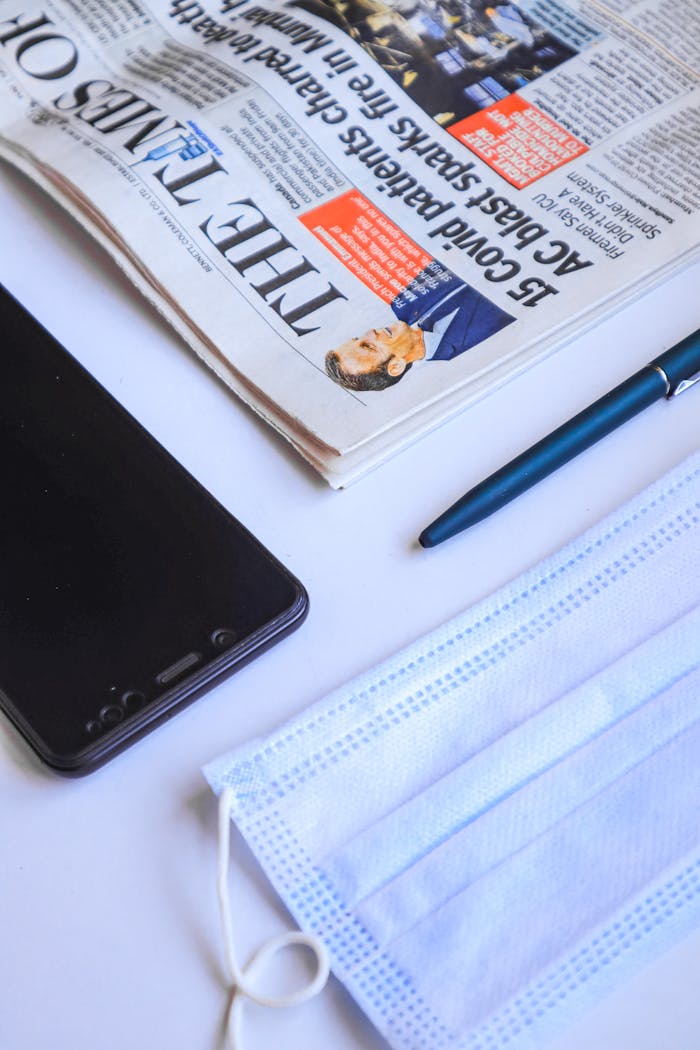 Flat lay of a newspaper, face mask, smartphone, and pen on a white background, capturing modern essentials.